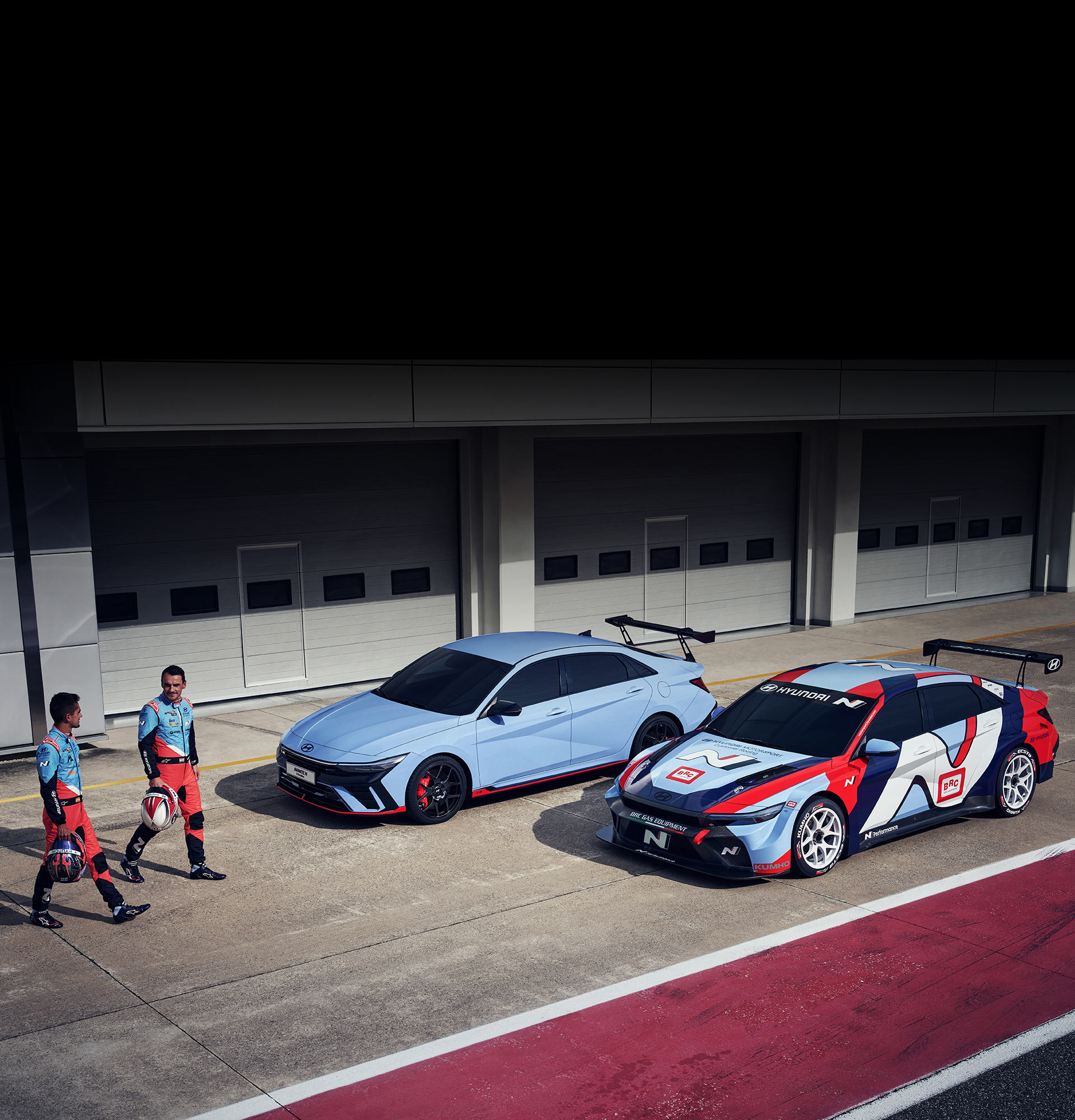 두대의 아반떼 N TCR 에디션 차량이 나란히 있는데, 우측의 차량에는 TCR 에디션 데칼이 장착되어있다. 나란히 서있는 차량 앞을 두명의 선수가 걷고 있는 사진.